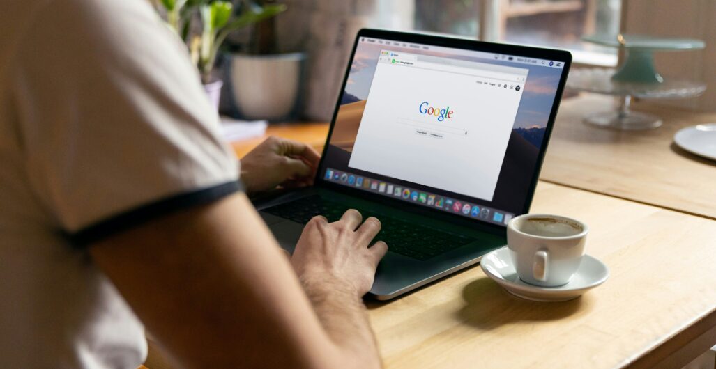 A person typing on a laptop with a Google search bar on the screen and a coffee cup in a saucer next to it.