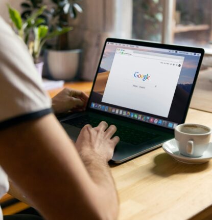 A person typing on a laptop with a Google search bar on the screen and a coffee cup in a saucer next to it.