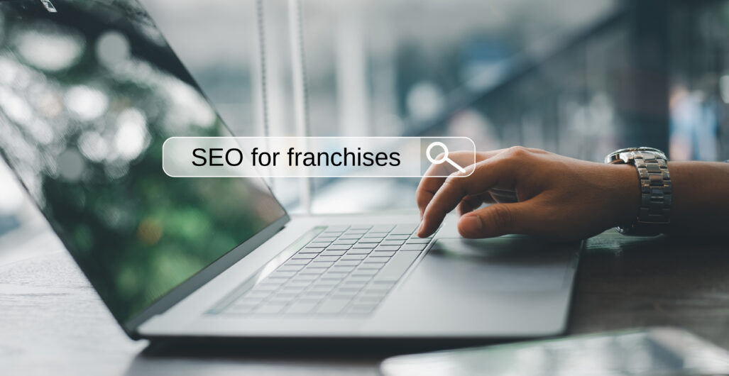 A hand types on a laptop keyword with a smartphone next to it and a graphic of a search bar is overlaid on the image with the text “SEO for franchises” in the search bar.