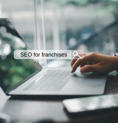 A hand types on a laptop keyword with a smartphone next to it and a graphic of a search bar is overlaid on the image with the text “SEO for franchises” in the search bar.