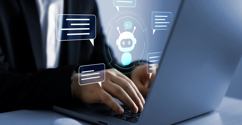 The torso of a person in a button-down dress shirt and sports jacket typing on a laptop with digital graphics showing chat bubbles surrounding a bot icon floating above the hands.
